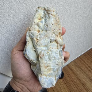 Puede incluir: Una roca grande, áspera, gris y blanca con cristales azules y blancos incrustados. La roca está siendo sostenida en la mano de una persona.