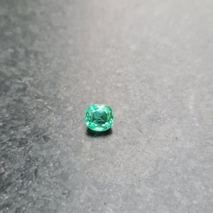 Puede incluir: Una esmeralda verde vibrante con talla coj&iacute;n, sobre un fondo gris.