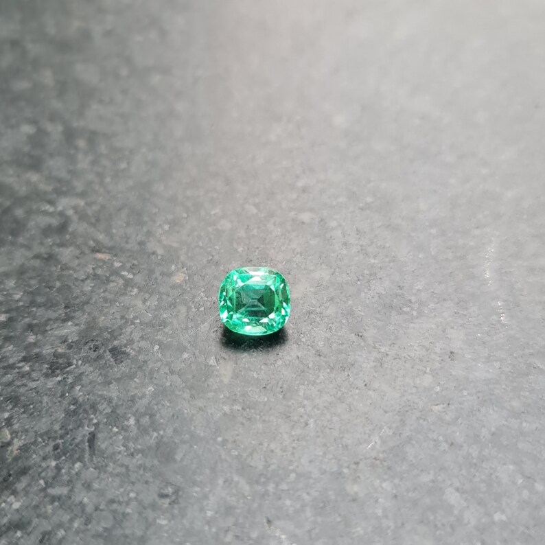 Puede incluir: Una esmeralda verde vibrante con talla coj&iacute;n, sobre un fondo gris.