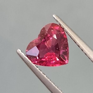 May include: A heart-shaped, faceted, deep pink gemstone is held by silver tweezers. The gemstone's facets reflect light, creating a sparkling effect. The background is a solid, light grey.