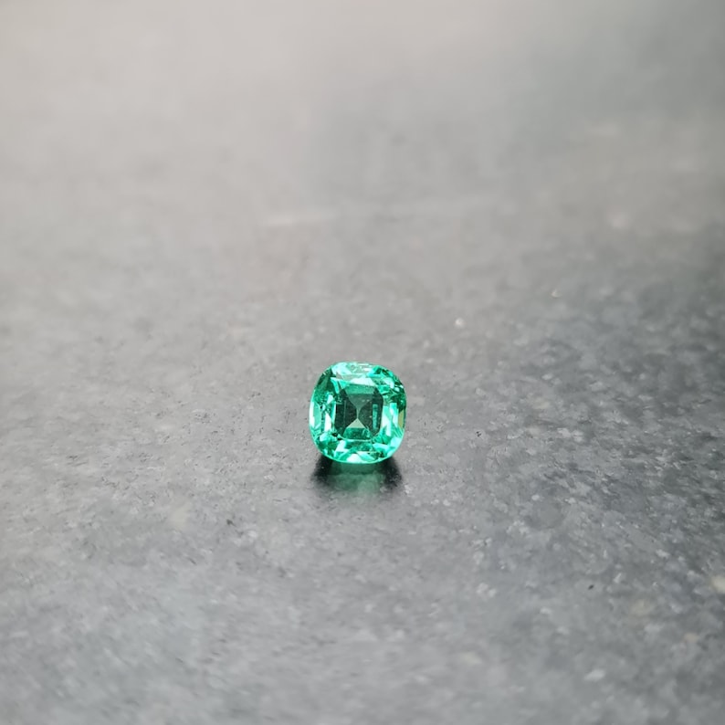 Puede incluir: Una gema de esmeralda verde vibrante con un corte de coj&iacute;n. La gema est&aacute; colocada sobre un fondo gris.