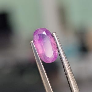 May include: A pink oval-shaped gemstone held by tweezers. The gemstone is faceted and has a light pink hue.