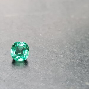 Puede incluir: Una esmeralda verde vibrante con un corte cuadrado de coj&iacute;n, sobre un fondo gris.