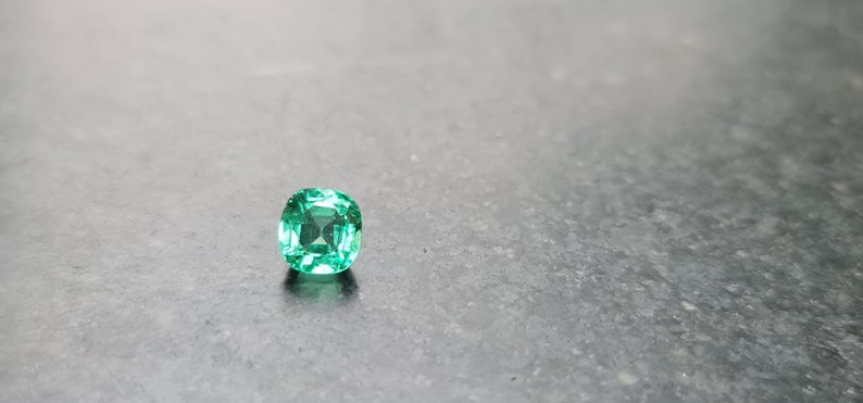 Puede incluir: Una esmeralda verde vibrante con un corte cuadrado de coj&iacute;n, sobre un fondo gris.