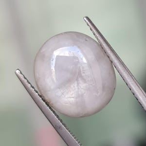 May include: A polished, oval-shaped, pale gray gemstone is held by a pair of silver tweezers. The gem reflects light, showcasing a smooth, rounded surface. The background is a soft, blurred green.