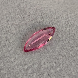 May include: A marquise-cut, pink gemstone. The gem is a vibrant shade of pink and reflects light, showcasing its internal structure. The stone is set against a grey background.