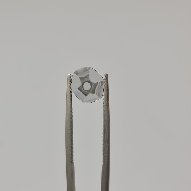 May include: A clear, faceted gemstone held by tweezers. The gemstone has a star-shaped pattern inside.