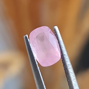 May include: A pale pink gemstone with a faceted cut, held in tweezers.