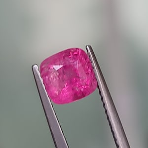 May include: A vibrant pink gemstone, likely a ruby, is held in tweezers. The gemstone is cut into a rectangular shape with rounded edges.