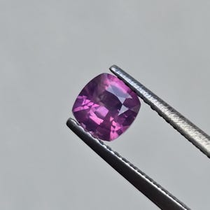 May include: A cushion-cut purple gemstone held in tweezers. The gemstone is a deep purple colour with a slight hint of pink.