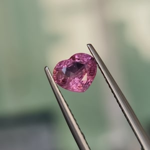 May include: A pink heart-shaped gemstone held in tweezers. The gemstone is faceted and has a light pink hue.