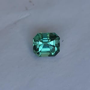 May include: A faceted, emerald-cut gemstone with a vibrant teal color. The gem has a rectangular shape with beveled edges, reflecting light and creating a sparkling effect. The gemstone is set against a white background.