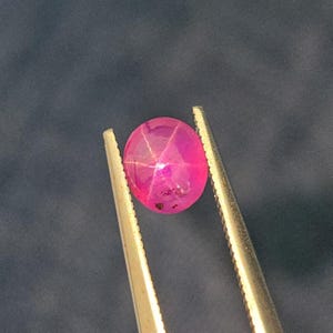 May include: A vibrant, oval-shaped, pink gemstone with a star-like pattern, held by tweezers against a dark background. The gem's surface reflects light, showcasing its internal structure and color variations. This gemstone is a potential jewelry component.