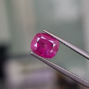 May include: A vibrant pink gemstone, likely a ruby, is held in a pair of tweezers. The gemstone is cut into a cushion shape and has a brilliant shine.