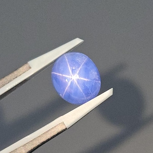 Op de afbeelding: Een blauwe ster-saffier edelsteen met een zespuntige ster in het midden. De edelsteen wordt vastgehouden door een pincet.