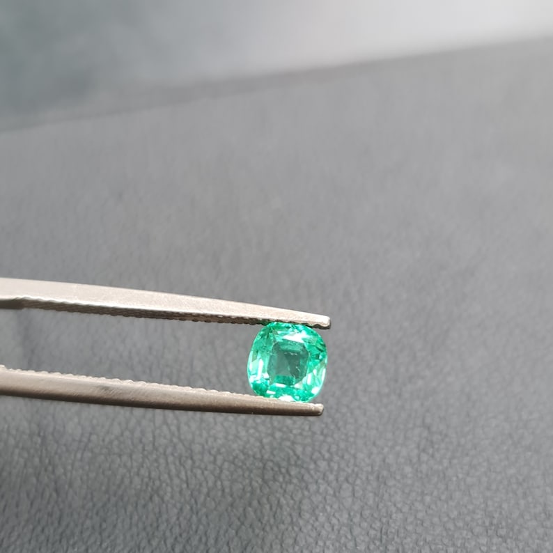 Puede incluir: Una esmeralda verde vibrante sostenida en unas pinzas. La esmeralda tiene un corte coj&iacute;n y es un hermoso ejemplo de esta piedra preciosa popular.
