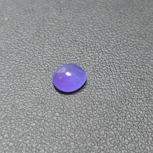 May include: A smooth, oval-shaped, purple gemstone. The polished stone has a glossy surface and is set against a textured, dark gray background. The gemstone is a vibrant shade of purple.