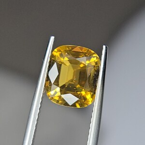 May include: A cushion-cut, golden-yellow gemstone held by silver tweezers. The gem has a faceted surface that reflects light, creating bright highlights. The overall shape is square with rounded corners, and the color is a rich, warm yellow.