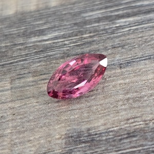 May include: A pink marquise cut gemstone with facets. The gemstone is on a wooden surface.