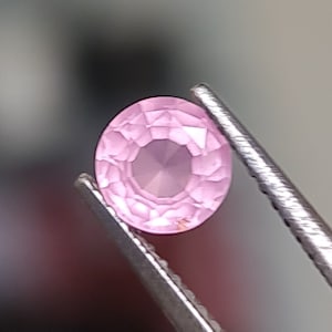 May include: A pink oval gemstone with a faceted cut, held in tweezers.