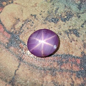 May include: Oval-shaped, purplish-pink gemstone with a six-rayed star pattern. The gem has a polished surface and a smooth, rounded shape. The star effect is created by light reflecting off of inclusions within the stone.