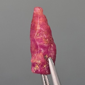 May include: A deep red ruby crystal with a pointed, elongated shape. The crystal has a rough, natural surface and is held by tweezers.