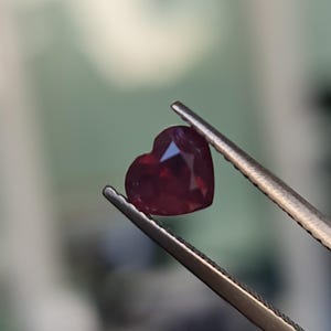 May include: A heart-shaped ruby gemstone held by tweezers. The ruby is a deep red colour and has a faceted cut.