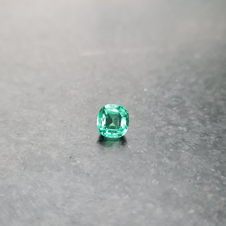 Puede incluir: Una gema de esmeralda verde vibrante con un corte de coj&iacute;n, sobre un fondo gris.
