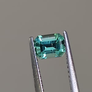 May include: A vibrant green gemstone with a rectangular emerald cut, held in tweezers.