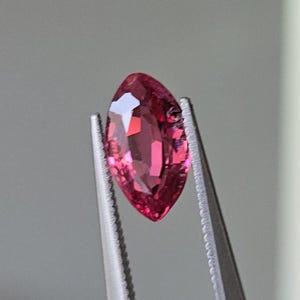 May include: A marquise-shaped pink gemstone held by tweezers. The gemstone is faceted and has a brilliant shine.
