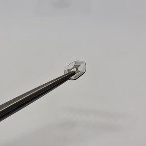 May include: A clear contact lens held by tweezers. The lens has a small hole in the center.
