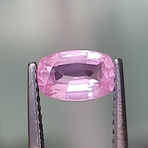 May include: A pink, oval-cut gemstone held by metal tweezers. The gem's facets catch the light, displaying a range of pink tones. The tweezers are silver, and the background is a soft, blurred green.
