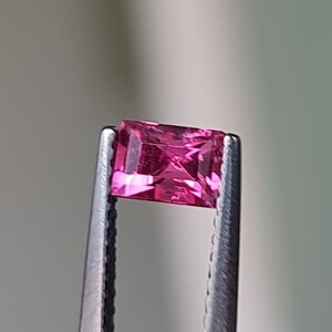 May include: A vibrant pink gemstone with a rectangular cut, held in place by metal tweezers.