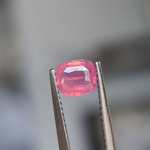 May include: A faceted, cushion-cut pink gemstone held by metal tweezers. The gemstone has a vibrant pink hue with visible internal structures and light reflections. The tweezers are silver-coloured, and the background is blurred.