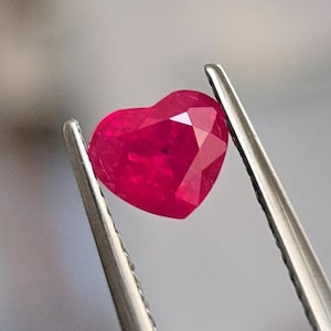 May include: A heart-shaped ruby gemstone with a deep red colour. The gemstone is held in tweezers.