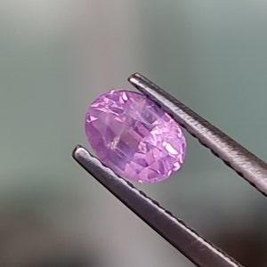 May include: A close-up of a pink oval gemstone held by tweezers. The gemstone has a faceted cut and a light purple hue.