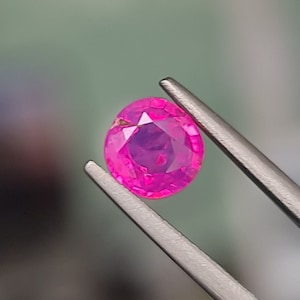 May include: A vibrant pink oval-shaped gemstone held by tweezers. The gemstone has a faceted cut and a slight purple hue.