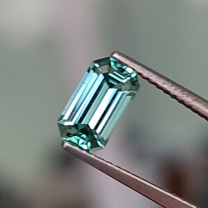 May include: A green gemstone with a rectangular emerald cut. The gemstone is held in tweezers.