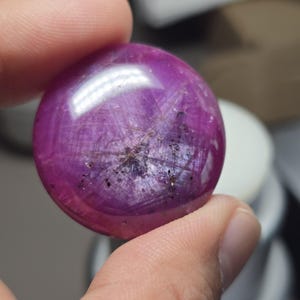 以下が含まれることがあります： 内部の筋と暗いインクルージョンを持つ、研磨された丸い濃いマゼンタ色の宝石。滑らかで反射する表面を示すように、宝石は指の間に保持されています。宝石はカボションです。