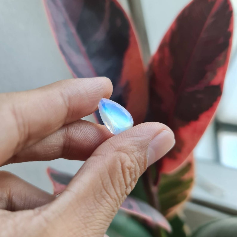 May include: A teardrop-shaped iridescent gemstone with a pearlescent sheen. The gemstone is held in a hand against a blurred background of a red and green leaf.