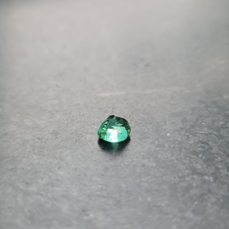 Puede incluir: Una gema de esmeralda verde vibrante con un corte en forma de coraz&oacute;n, mostrada sobre una superficie gris.