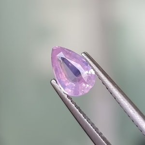 May include: A pear-shaped pink sapphire gemstone held by tweezers. The gemstone is translucent and has a light purple hue.