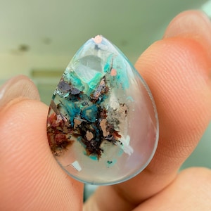 Peut inclure: Un pendentif en forme de goutte d'eau transparent. L'intérieur présente un mélange de couleurs, notamment du turquoise, du marron et du rouge, créant un motif abstrait unique. Le pendentif est tenu entre les doigts, mettant en valeur sa forme et sa taille.