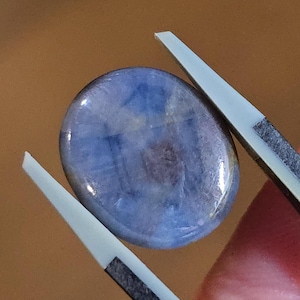 May include: A blue oval gemstone with a smooth, polished surface. The gemstone is held in place by two tweezers.