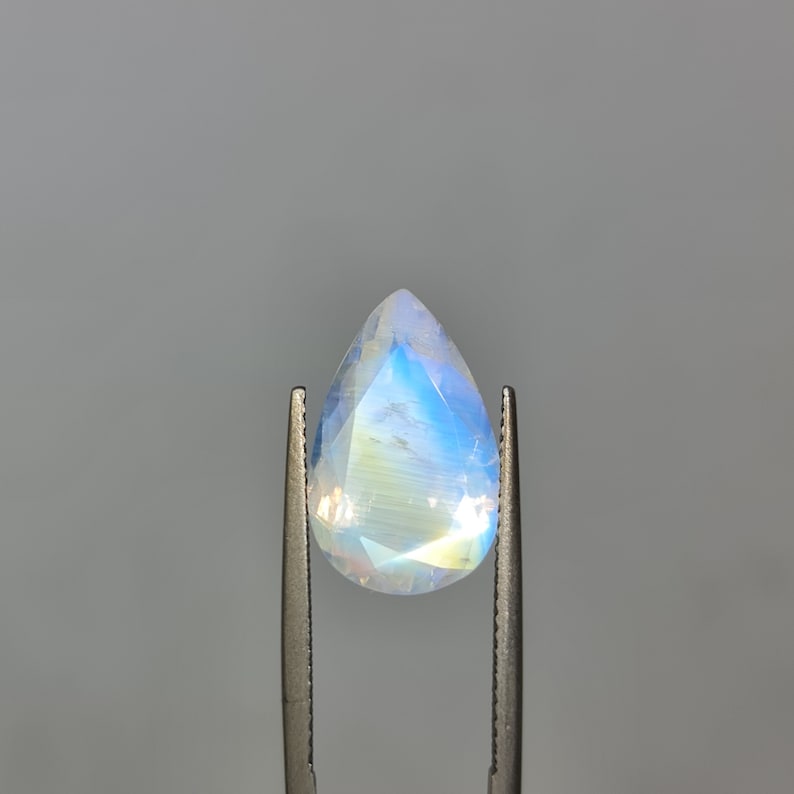 May include: A pear-shaped moonstone gemstone with a blue iridescent sheen, held in tweezers.