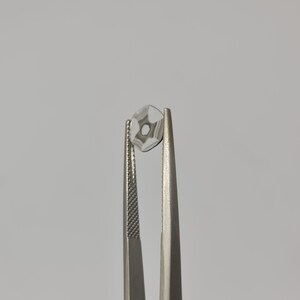 May include: A clear glass lens with a star-shaped pattern is held by a pair of silver tweezers.