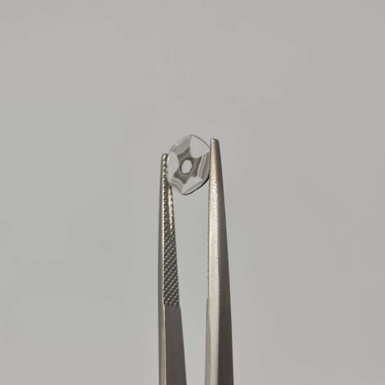 May include: A clear glass lens with a star-shaped pattern is held by a pair of silver tweezers.