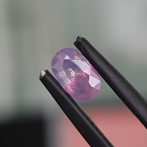 May include: A light purple oval-shaped gemstone held by tweezers.
