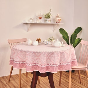 May include: A round pink tablecloth with a white damask pattern covers a dark wooden table. The table is set with a teapot, creamer, sugar bowl, and a bowl of cookies. Two pink chairs flank the table.