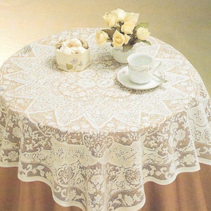 May include: A round table covered with a white lace tablecloth. The tablecloth has a floral design and a scalloped edge. A small vase of yellow roses, a teacup and saucer, and a small decorative box are on the table.